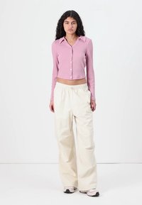 Rosa ribbad kort cardigan med krage och knappstängning, kombinerad med löst sittande krämfärgade byxor. Modellen står framför en enfärgad bakgrund.