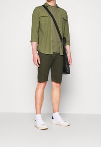 Chemise olive à manches longues avec poches poitrine, shorts assortis et baskets blanches. Comprend un sac bandoulière noir. Texture lisse.