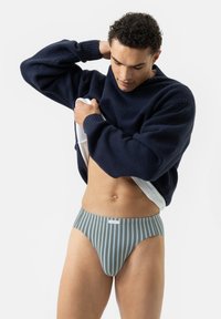 Mann hebt einen marineblauen Pullover an, trägt grün und grau gestreifte Unterwäsche mit vertikalen Streifen und steht vor einem schlichten weißen Hintergrund.