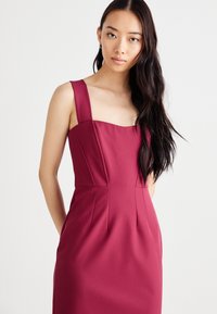Burgundská priliehajúca šaty s hrubými štvorcovými ramienkami, vystuženým telom a skladmi. Vyrobené z hladkej tkaniny s elegantným strihom.
