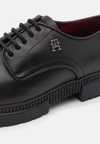 Chaussure en cuir noir avec une finition texturée, conçue avec un laçage, un petit détail de logo, et une semelle noire rainurée pour une meilleure adhérence.