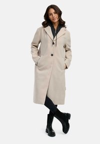 Beige lange jas met een op maat gemaakt ontwerp, voorzakken en zwarte knopen, gedragen over een donkere hoodie en gecombineerd met zwarte boots met een dikke zool.