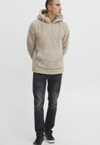 Beige fleece pullover with a hood, front half-zip, and kangaroo pocket. Worn with dark jeans and black sneakers, featuring a textured fabric.