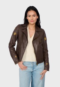 Veste de moto en cuir marron avec des accents dorés en forme d'étoile sur les manches, fermetures éclair et col à revers, associée à un haut en tricot crème.