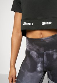 Camiseta negra recortada con "STRONGER" impreso dos veces en texto blanco. Leggings de cintura alta con un patrón oscuro y nublado y paneles de malla.