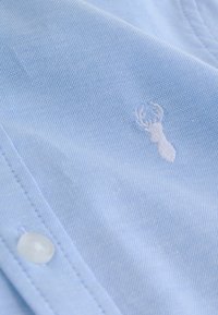 Chemise en tissu texturé bleu clair avec tête de cerf et bois brodés blancs près d'un bouton blanc et d'une couture cousue.