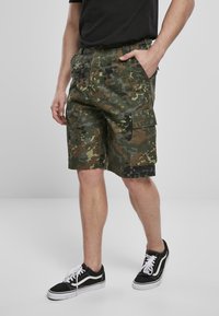 Pantalones cortos de camuflaje en verde con acentos en negro, con múltiples bolsillos y un corte relajado, combinados con zapatillas negras y blancas.