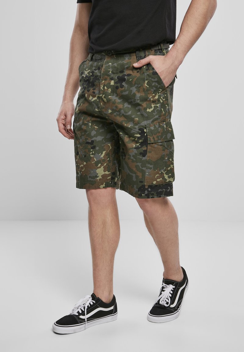 Pantalones cortos de camuflaje en verde con acentos en negro, con múltiples bolsillos y un corte relajado, combinados con zapatillas negras y blancas.