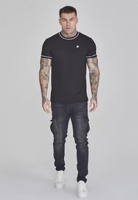 Juoda marškinėliai su apvalia iškirpte ir balta apvada, derinami su tamsiomis kroso stiliaus džinsais su šoniniais kišenėmis ir baltais sportbačiais.