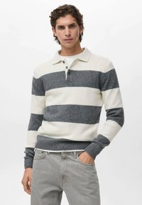 Joven con cabello corto castaño, vistiendo un suéter de rayas anchas en blanco y gris y jeans gris claro, con una mano en el bolsillo.