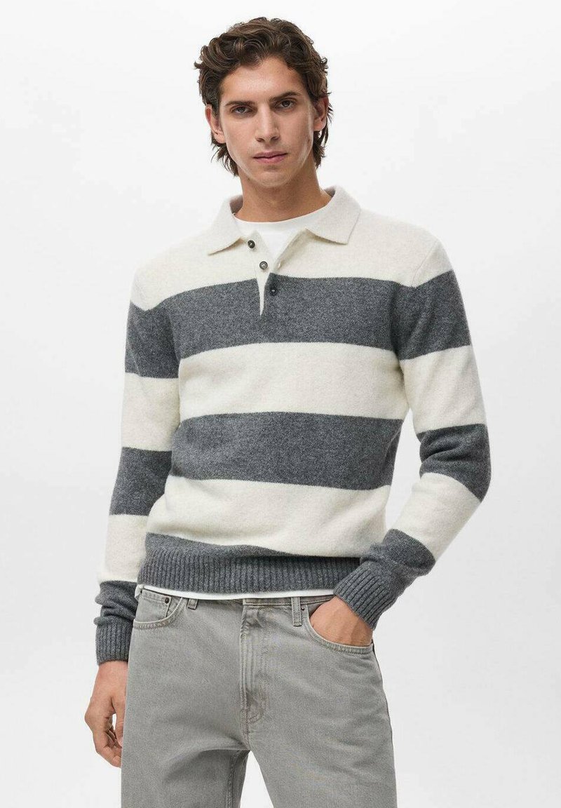 Joven con cabello corto castaño, vistiendo un suéter de rayas anchas en blanco y gris y jeans gris claro, con una mano en el bolsillo.