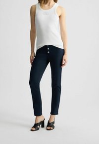Camiseta sin mangas blanca con textura acanalada y logo, combinada con pantalones slim-fit azul marino con cierre de cinco botones y sandalias de tacón negras.