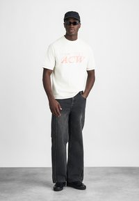 White cotton t-shirt with red printed logo, paired with loose-fitting black jeans. Accessories include a black cap and dark sunglasses.