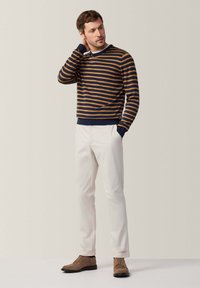 Maglione a righe blu navy e senape, pantaloni bianchi e scarpe in suede marroni. Il maglione presenta polsini e orlo a costine, aderendo strettamente in vita.