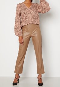 Blusa marrone con stampa floreale, scollatura a V e lunghe maniche a sbuffo, abbinata a pantaloni in ecopelle beige e scarpe nere con tacco.