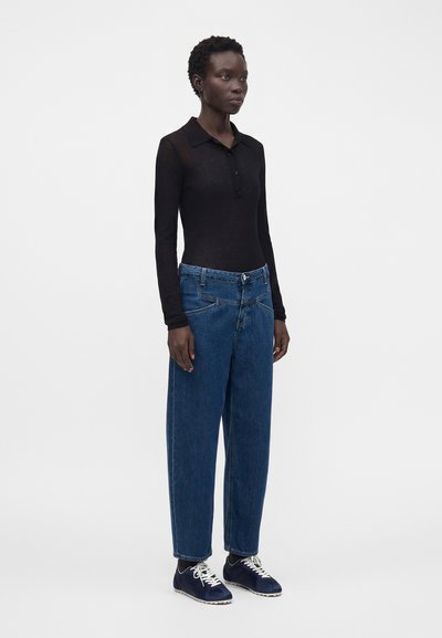 Personne portant un polo noir cintré à manches longues, un jean bleu large et des baskets bleu marine, debout devant un fond blanc uni.