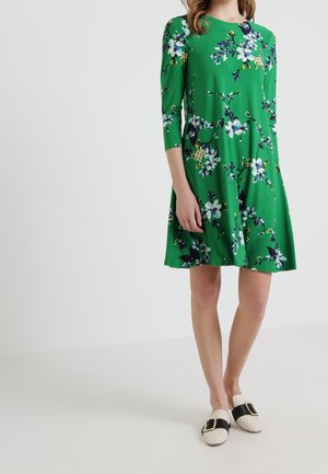 Robe florale verte à manches trois-quarts, arborant des fleurs blanches et bleues, jupe évasée, tissu doux, associée à des chaussures blanches à enfiler avec des accents noirs.