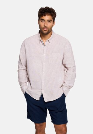 Hombre con cabello rizado que lleva una camisa beige claro de botones con bolsillo delantero y pantalones cortos azul oscuro, de pie con las manos en los bolsillos.