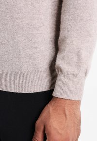 Ljusgrå tröja i mjukt stickat tyg, med ribbad nederkant och ärmslut. Närbild av ärmen visar struktur och passform.