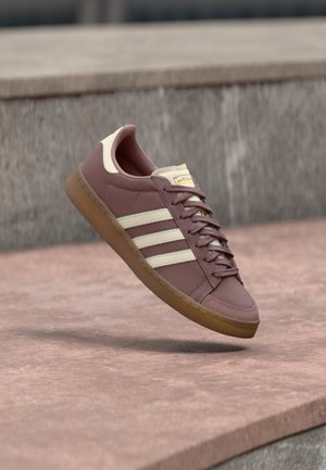 Zapatilla de cuero rosa con detalles en beige, con tres franjas blancas, punta redonda, suela de goma marrón y cordones. Diseño clásico con detalles de costura.