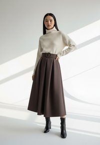 Kremowy sweter z prążkowanym golfem w połączeniu z brązową plisowaną spódnicą midi z okrągłą klamrą paska, uzupełniony czarnymi botkami za kostkę.