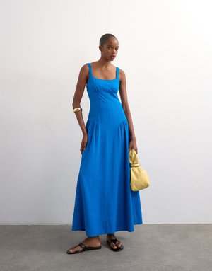 Kvinde iført en blå maxikjole uden ærmer, sorte sandaler, guldarmbånd og med en gul håndtaske foran en ensfarvet hvid væg.