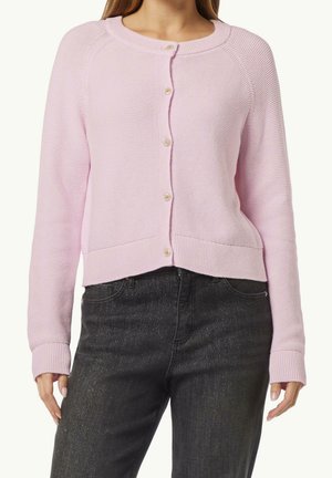 Femme portant un cardigan en maille rose clair boutonné avec des manches longues et un jean taille haute noir avec de discrètes paillettes.