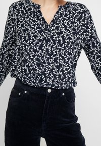 Žena v námořnickém blůzce s bílým vzorem bobulí, zasunutou do kalhot z černého kordu s vysokým pasem, na jednolitém pozadí.