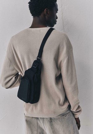 Bărbat purtând un pulover bej și blugi gri, care poartă o geantă neagră mică pe umărul drept, în fața unui perete simplu.