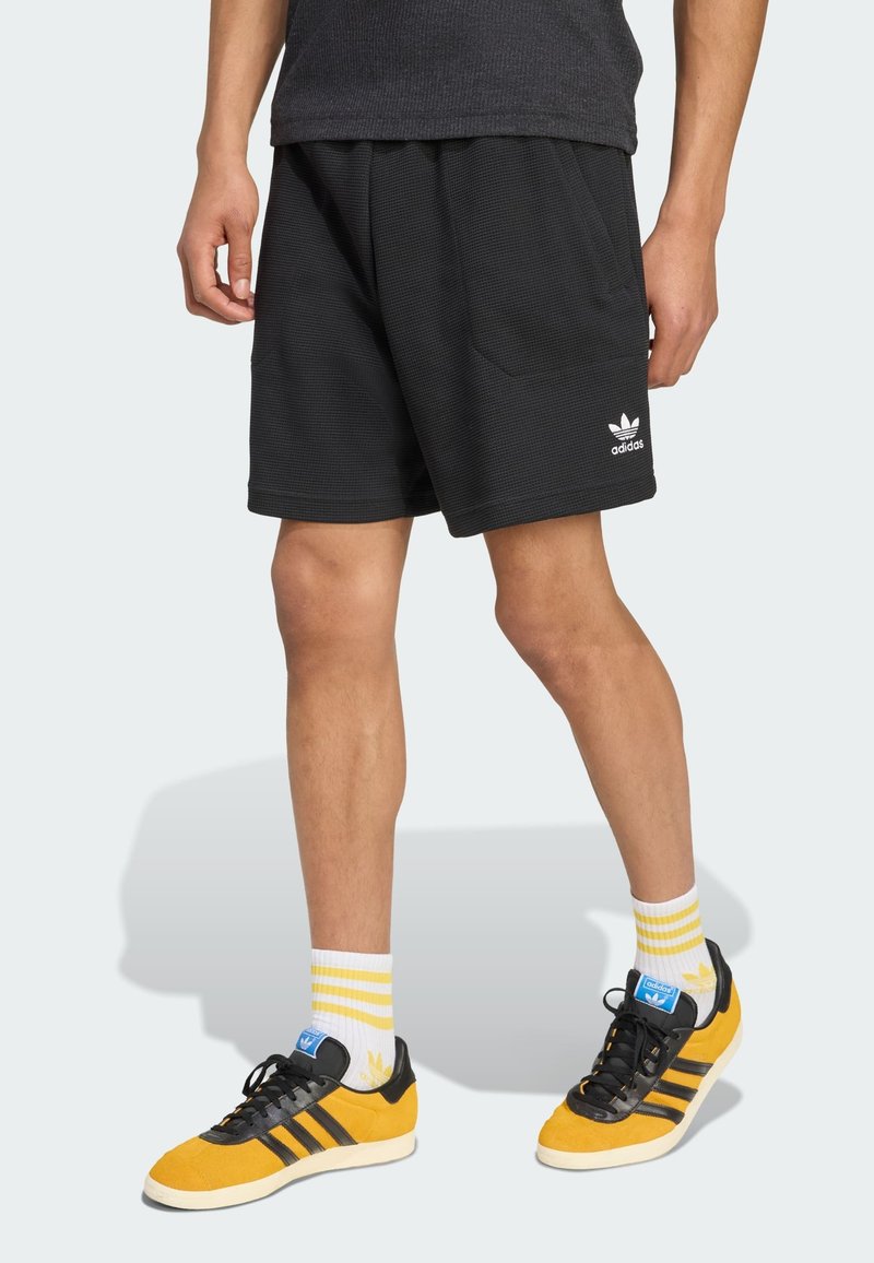 Shorts neri a trama con una vestibilità ampia, dotati di tasche laterali e un logo Adidas bianco sull'orlo, abbinati a scarpe gialle e calze a righe.