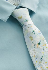 Lys blå skjorte med krave i et struktureret stof, parret med et mønstret slips i pastelfarver med blomsterdesign og grønne accenter.