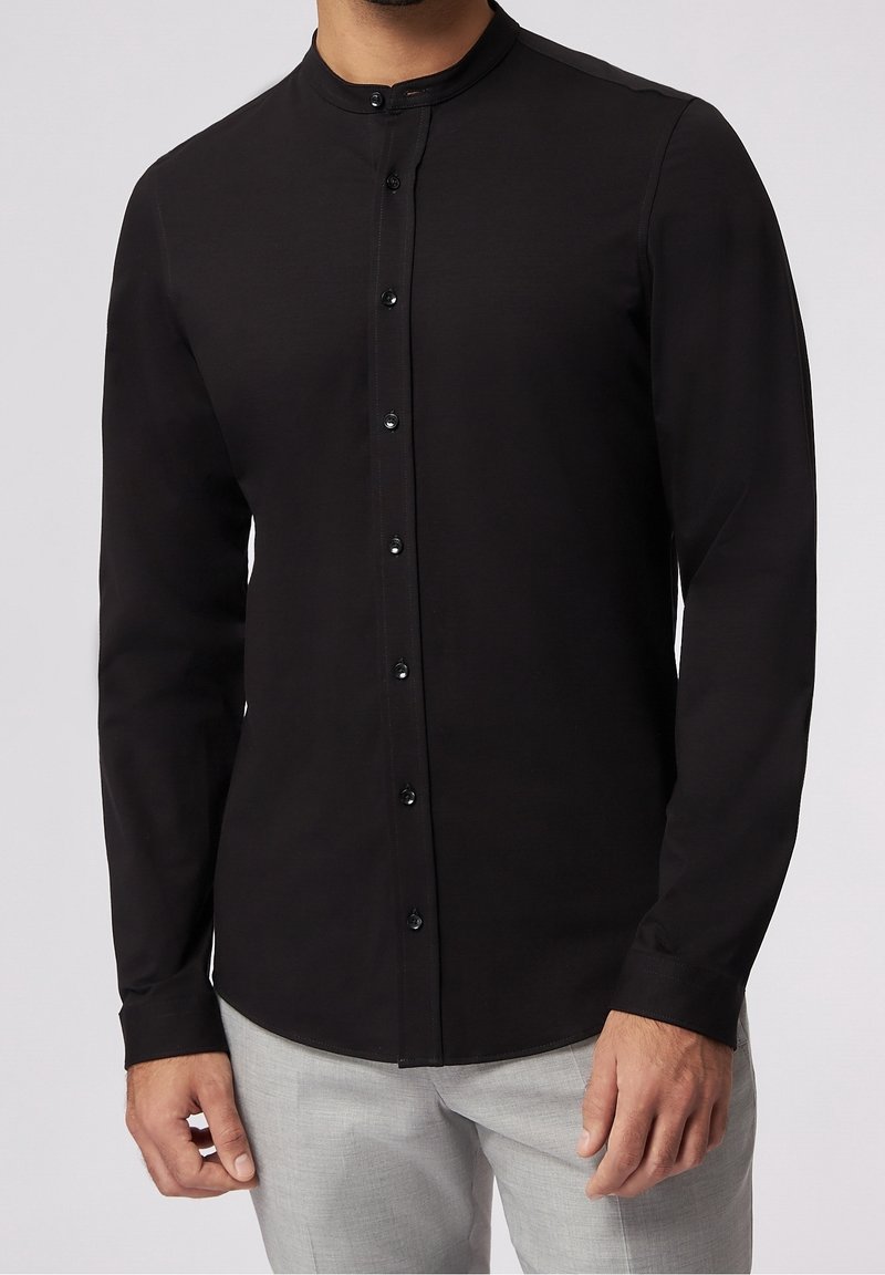Camisa negra de manga larga con cuello de banda y placket de botones en el frente. Presenta una textura suave y un diseño ajustado. Detalles simples en toda la prenda.