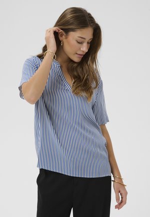 Tânără atingându-și părul, purtând o bluză cu dungi verticale albastre și albe, pantaloni negri și bijuterii aurii, pe un fundal simplu.