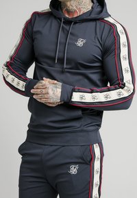 Morska granatowa bluza sportowa z czerwono-białymi bocznymi paskami, logo na piersi, kapturem ze sznurkiem i dopasowanym krojem. Wykonana z gładkiego, elastycznego materiału.