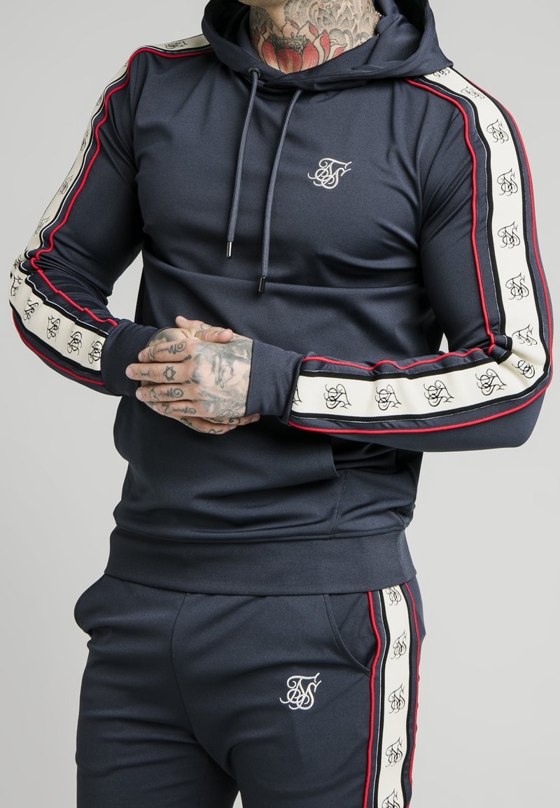 Morska granatowa bluza sportowa z czerwono-białymi bocznymi paskami, logo na piersi, kapturem ze sznurkiem i dopasowanym krojem. Wykonana z gładkiego, elastycznego materiału.