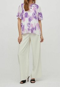 Vit blus med lila blommönster, volangärmar, tillsammans med högmidjade vita byxor och svarta sandaler med öppen tå.