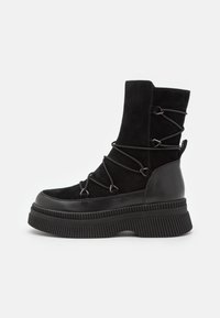 Zwarte enkellaars met een bovenwerk van suède, veterontwerp met metalen accenten, en een dikke ribbelzool voor extra hoogte en ondersteuning.
