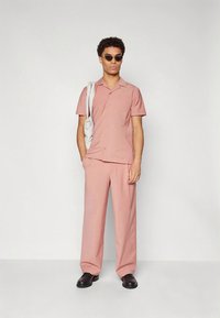 Rosa kortermet skjorte med knappelukking, matchende rosa bukser og svarte sko. Modellen bærer en hvit veske og har på seg solbriller.