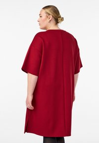 Červené voľné šaty s krátkymi rukávmi, uvoľneným strihom, bočnými rozparkami a hladkou textúrou. Viditeľný šev na zadnej strane.