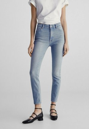 Jean slim fit bleu clair porté avec un t-shirt blanc aux manches retroussées et des chaussures noires à talons bas avec bride à la cheville sur un fond uni.