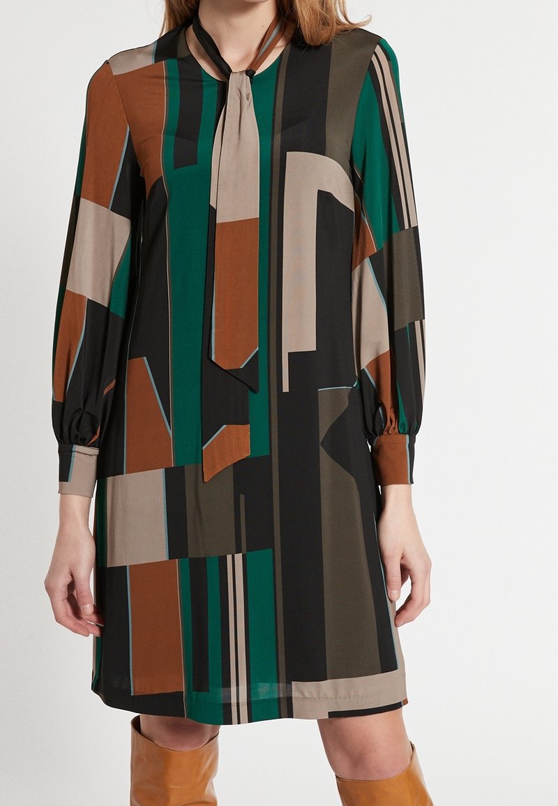 Robe à manches longues avec un motif géométrique multicolore présentant du vert, du marron, du noir et du beige. Tissu doux avec un détail de lien au cou et des poignets froncés.