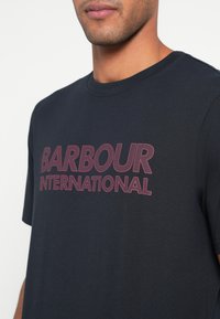 Tume sinine puuvillane T-särk, millel on esiküljel julge, mustriline "BARBOUR INTERNATIONAL" logo roosas värvitoonis. Lühikesed varrukad ja ümmargune kaelus.