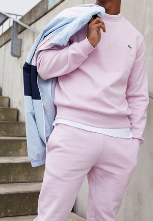 Sudadera rosa claro y pantalones a juego, hechos de tela suave. Lleva una chaqueta bomber azul y marina con cremallera y detalles de malla.