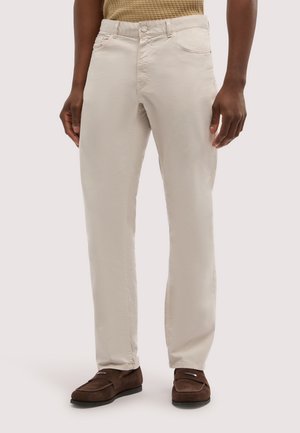 Uomo che indossa pantaloni beige a gamba dritta, mocassini in suede marrone scuro e una maglia testurizzata color senape, in piedi davanti a uno sfondo bianco.