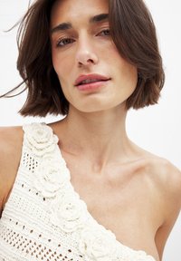 Valge heegeldatud ülaosa, mis omab ühte õlga katvat disaini, tekstuursete lillede aktsentidega rihmal ning delikaatse, avatud kootud mustriga.