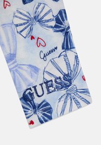 Tissu blanc orné de motifs de nœuds bleus et rouges, avec un logo "GUESS" bleu texturé et de petits cœurs rouges. Texture lisse, motif ludique.