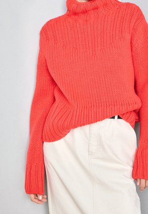 Woman wearing a thick, bright orange knitted sweater tucked into a high-waisted off-white skirt, standing against a plain background.