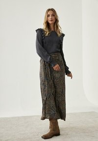 Suéter gris de manga larga con hombros acampanados, combinado con una falda maxi estampada que presenta motivos de hojas en colores oscuros. Botas marrones hasta el tobillo.