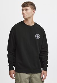 Svart hettegenser med rund hals, lange ermer og ribbede mansjetter. Har et hvitt grafisk design på venstre brystområde. Myk tekstur.