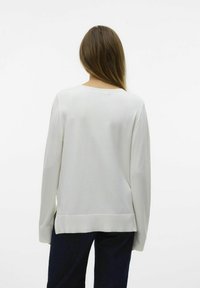 Haut beige clair à manches longues avec un col rond, présentant une texture douce et des fentes sur les côtés, associé à un pantalon en denim foncé.